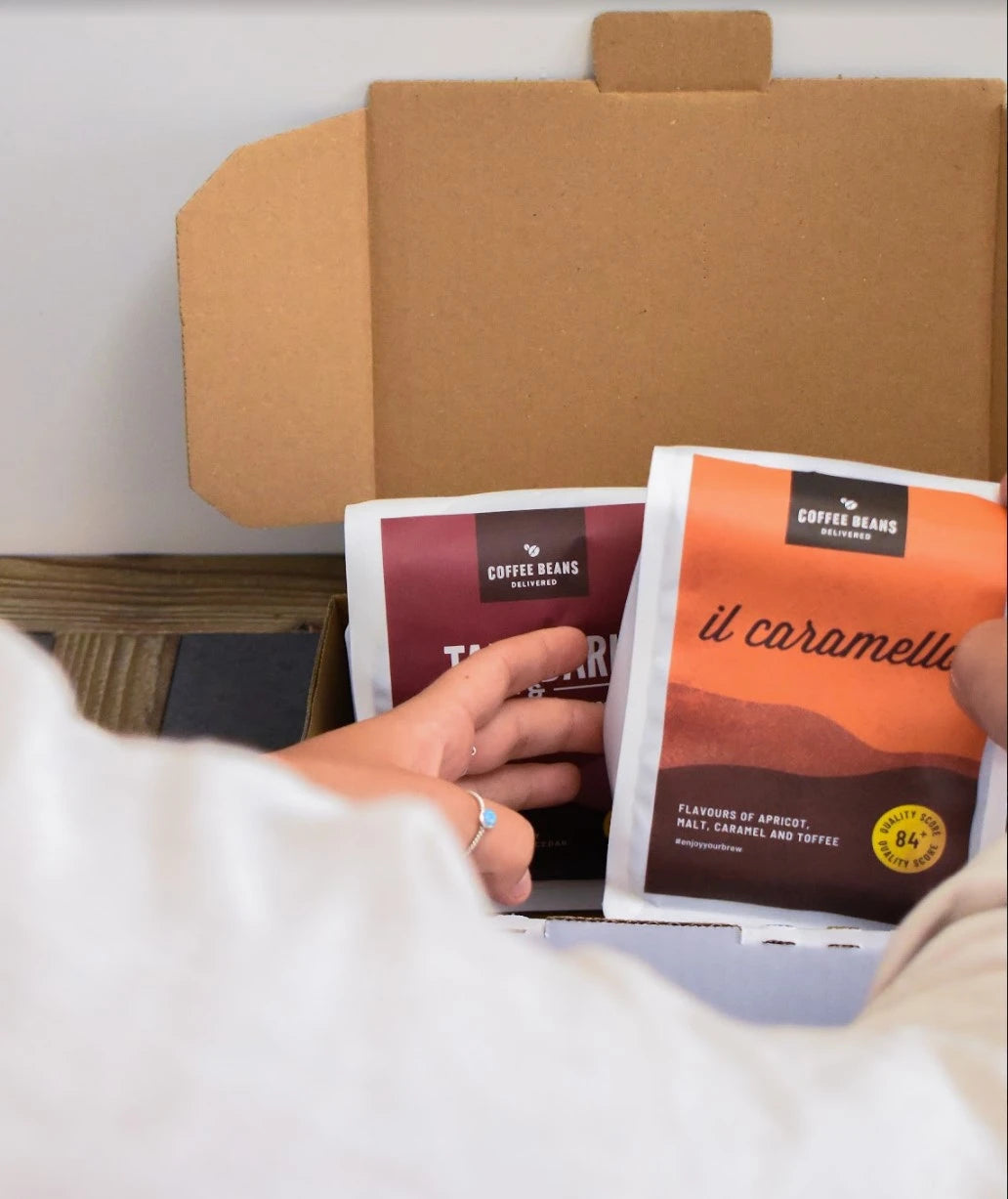 Two bags of coffee beans of different blends sit in a box while a person reaches in and grabs a bag of il caramello organic coffee beans
