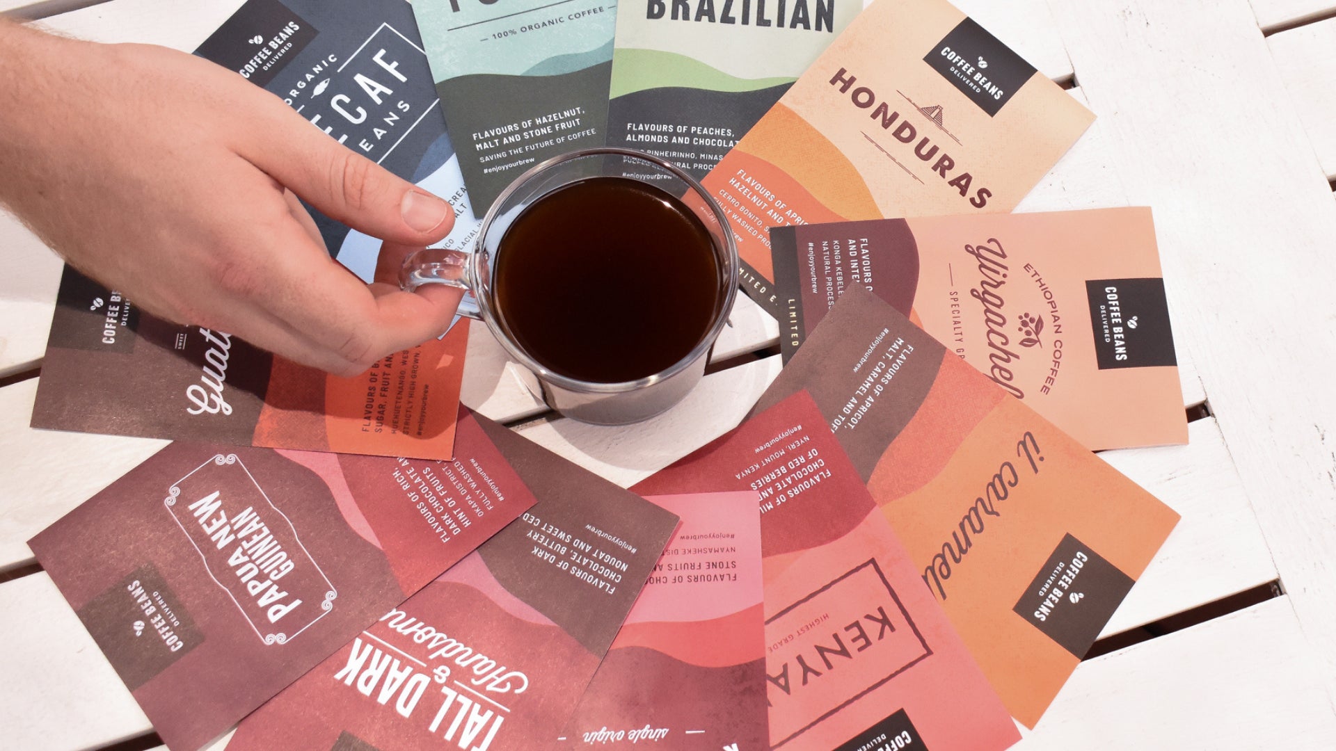 A black espresso coffee sits in the middle of a circle of colourful coffee single origin labels with a male hand reaching for the coffee